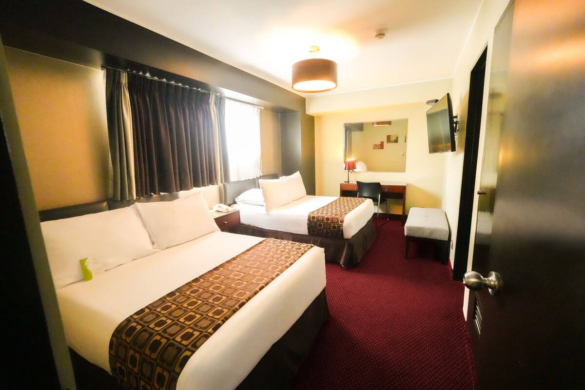 Quarto de hotel com duas camas, uma escrivaninha, uma cadeira, uma mesinha, um espelho e um tapete vermelho.