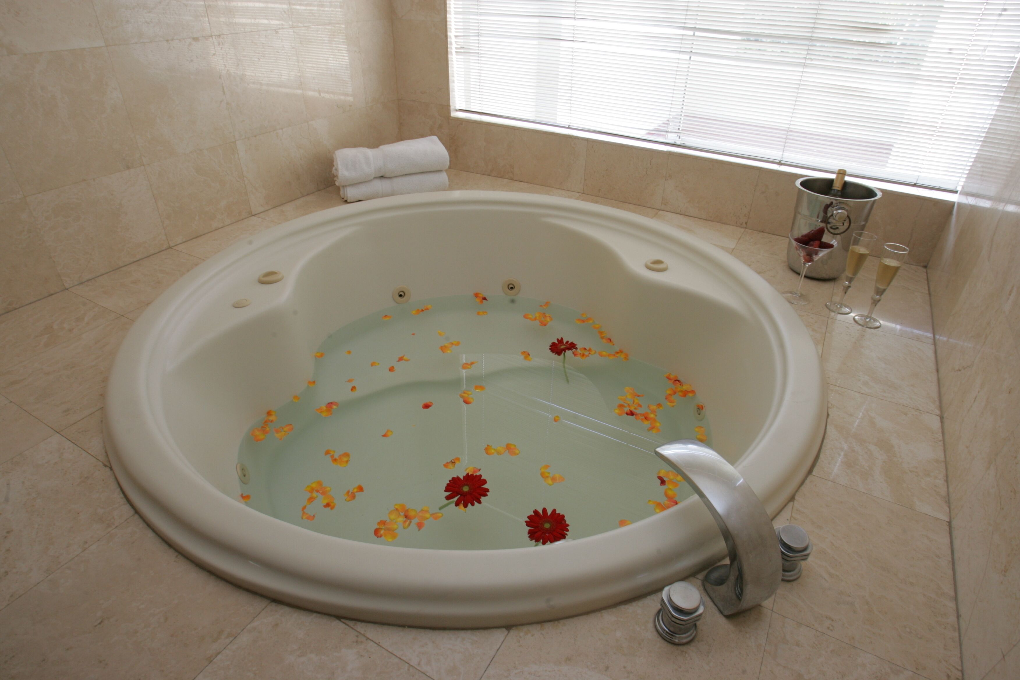 A large bathtub with flower petals floating on the water, a towel, and a champagne bottle with glasses on the floor.