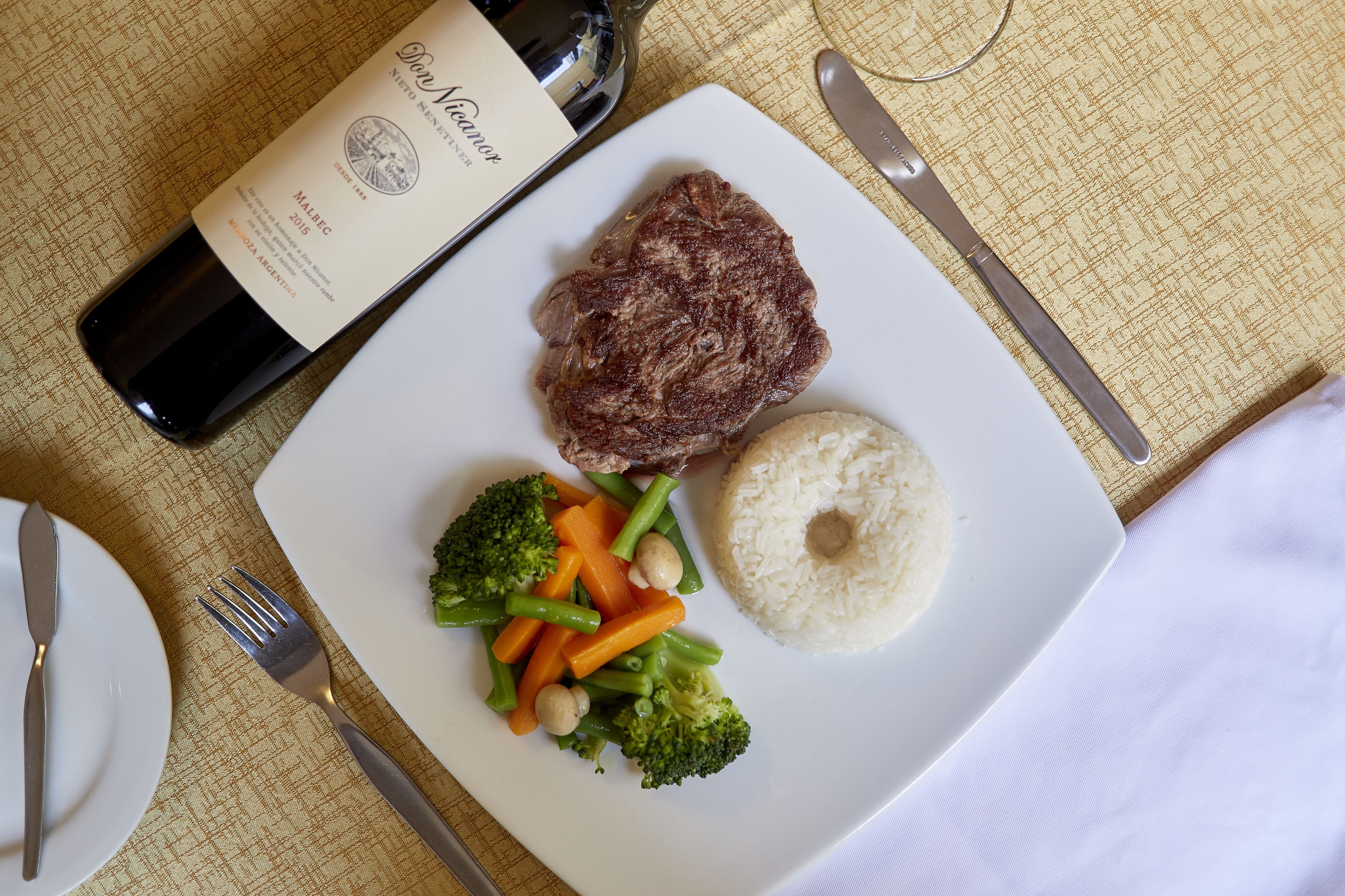 Un plato con carne, arroz y verduras, una botella de vino y cubiertos sobre un mantel.
