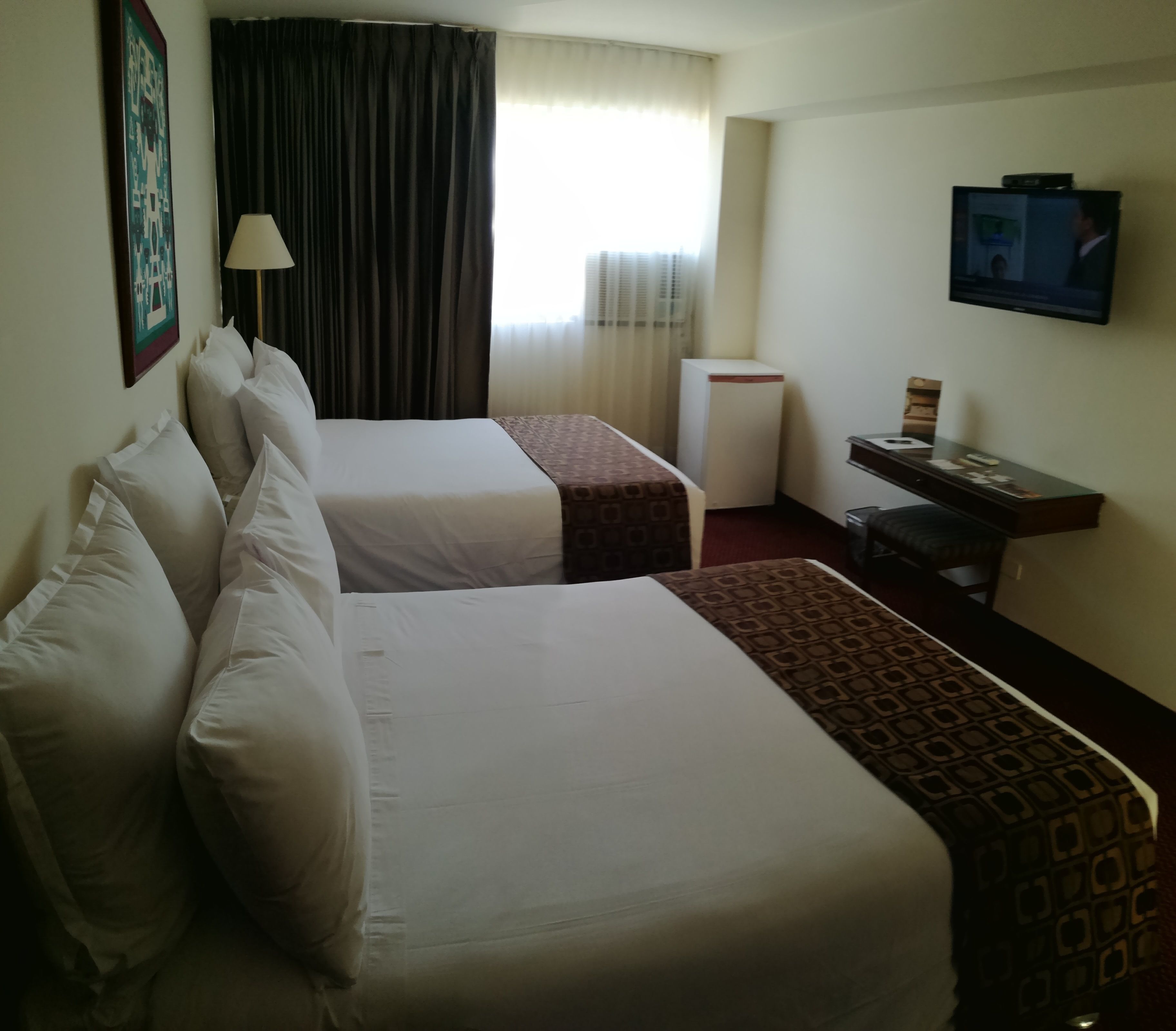 Habitación de hotel con dos camas, un escritorio, televisor y ventana con cortinas.