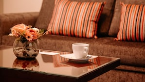Une table basse en verre, sur laquelle se trouvent une tasse et une soucoupe blanches, un vase avec des fleurs et des coussins à rayures sur un canapé marron.