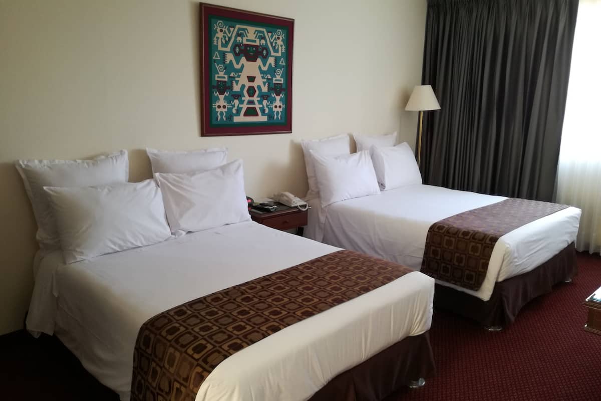 Duas camas de casal com roupa de cama branca e cabeceiras marrons, uma mesa de cabeceira com telefone, abajur e um quadro na parede.