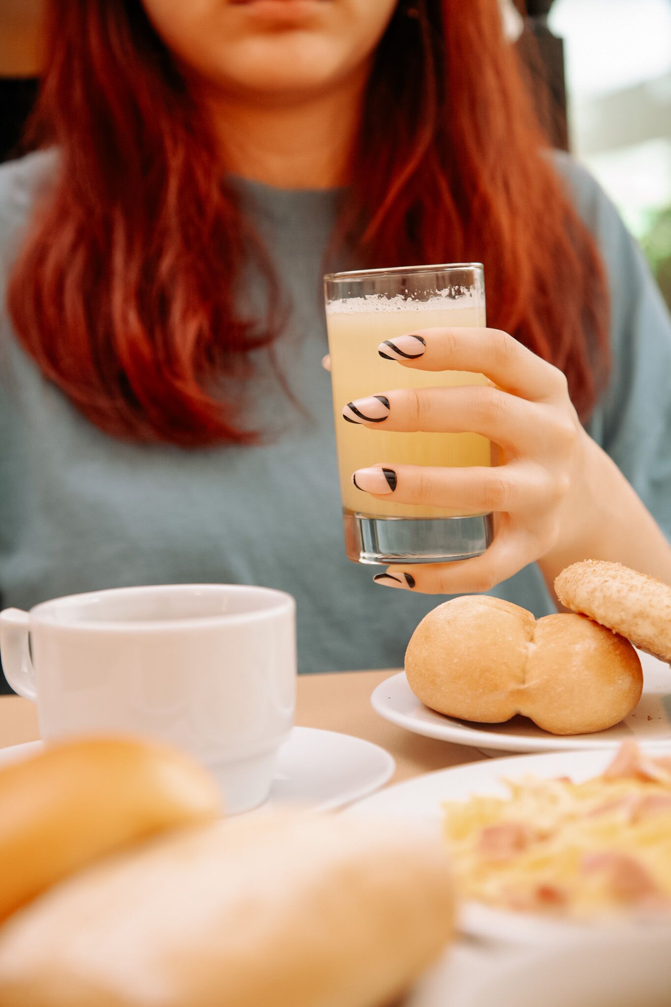 A person holding a glass of a beverage, with a white mug and food items on the table.