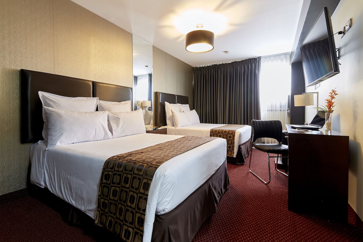 Quarto de hotel com duas camas, uma mesa, uma cadeira, uma televisão e uma janela grande com cortinas.