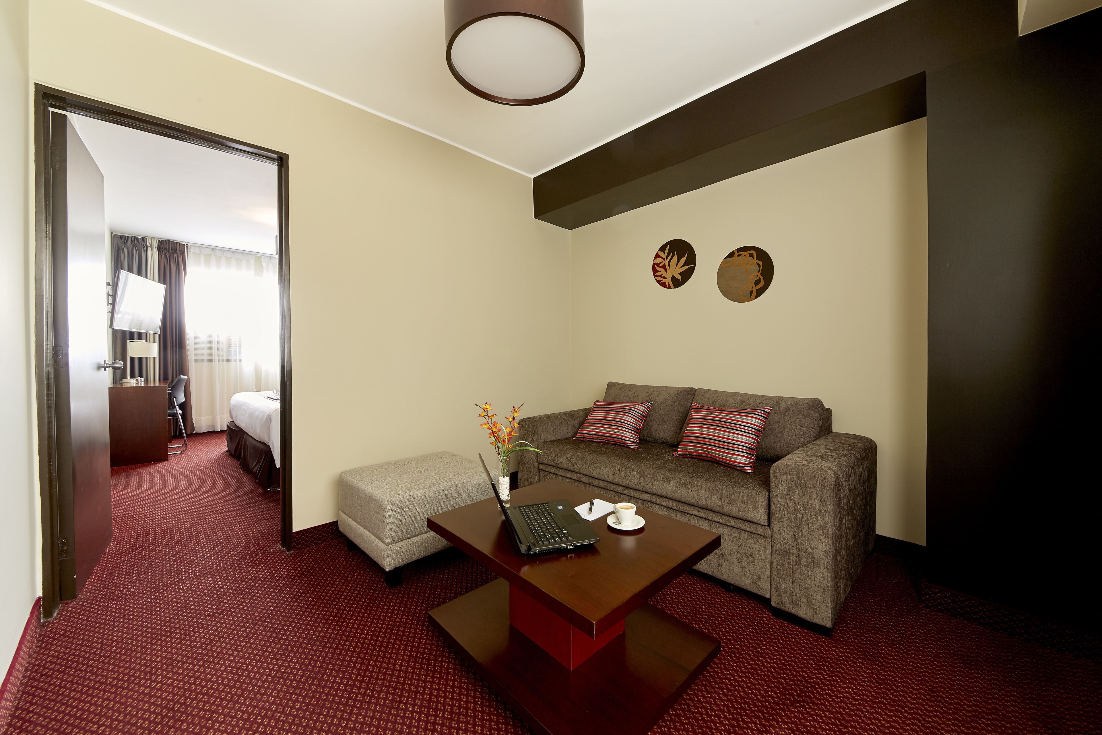 A hotel room with a red carpet, a grey sofa, a wooden coffee table with a laptop and a cup, and a bed in the background.