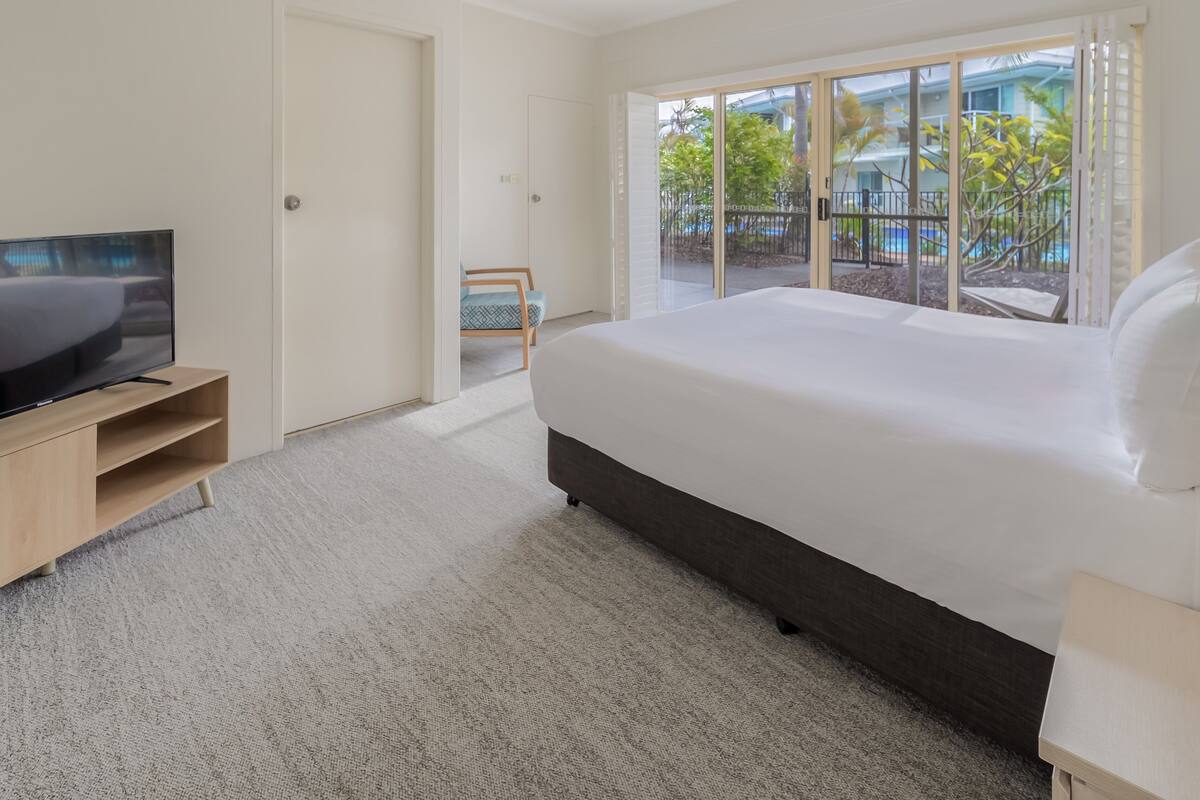 A modern hotel room with a large bed, a TV on a wooden stand, and a view of a pool and greenery through large windows.