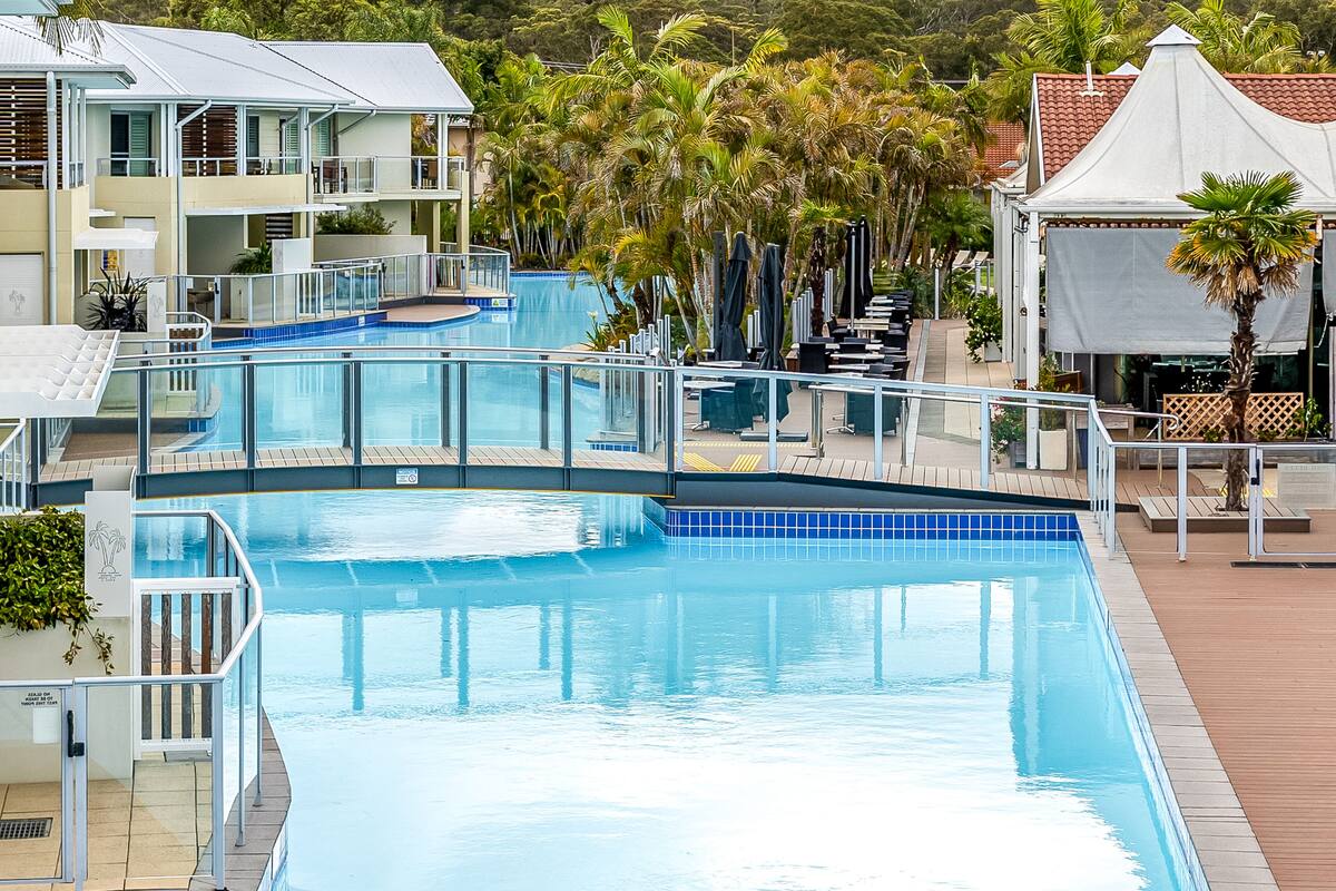 A large, clear swimming pool with a wooden deck surrounding it, a covered seating area, and a view of a building with multiple balconies.