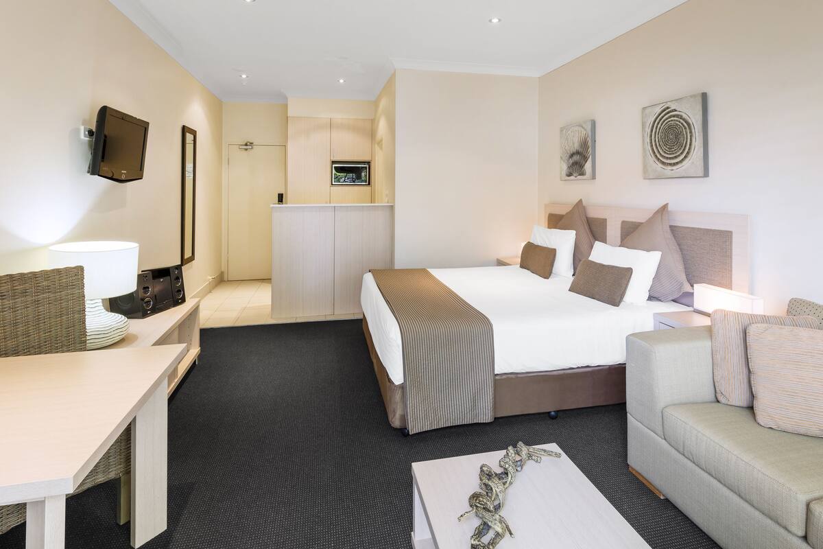 A hotel room with a bed, a sofa, a desk, a TV, and a kitchenette.