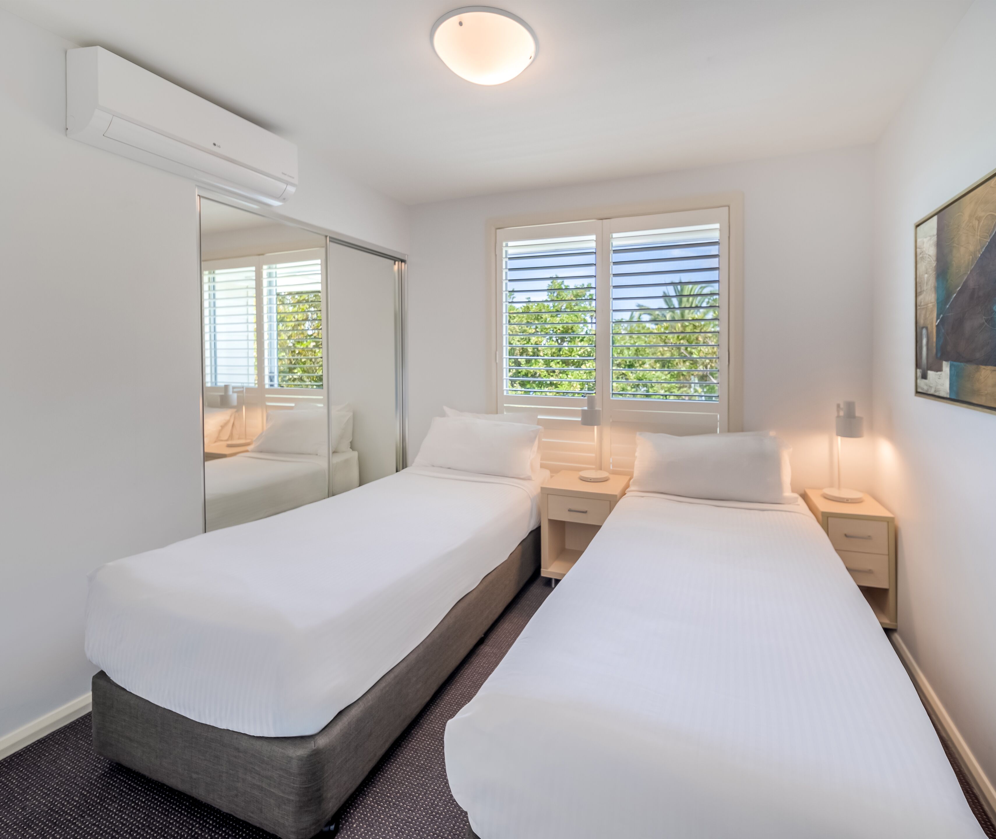 A hotel room with two single beds, a large mirror, a window with blinds, and a painting on the wall.