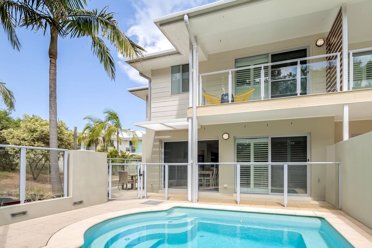 A modern two-story house with a swimming pool, large windows, and a balcony with yellow chairs.