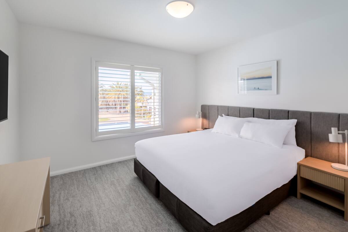 A modern hotel room with a large bed, a nightstand, a desk, and a window with blinds.