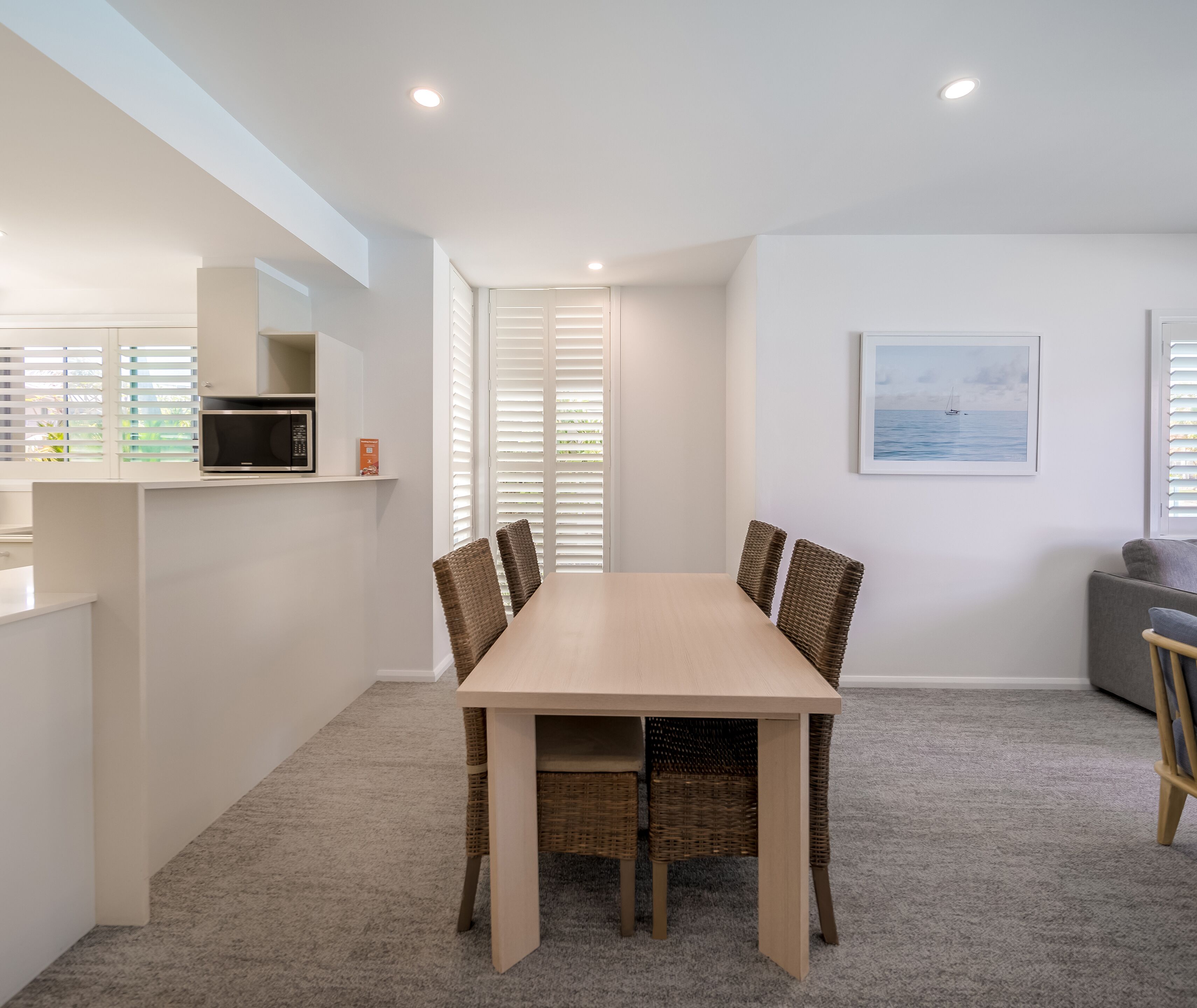 Una cocina moderna con armarios blancos, microondas y comedor con mesa de madera clara y sillas de mimbre.