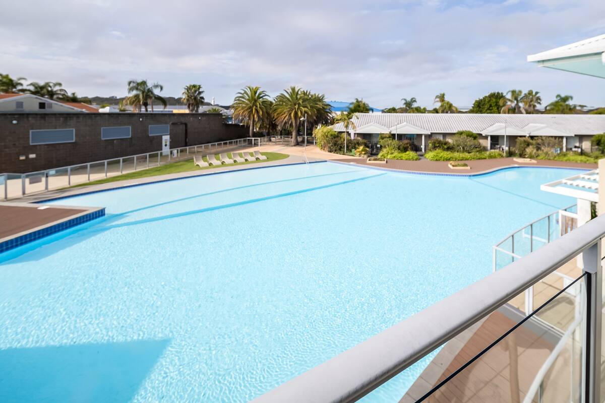 A swimming pool with lounge chairs, a covered area, and palm trees in the background.