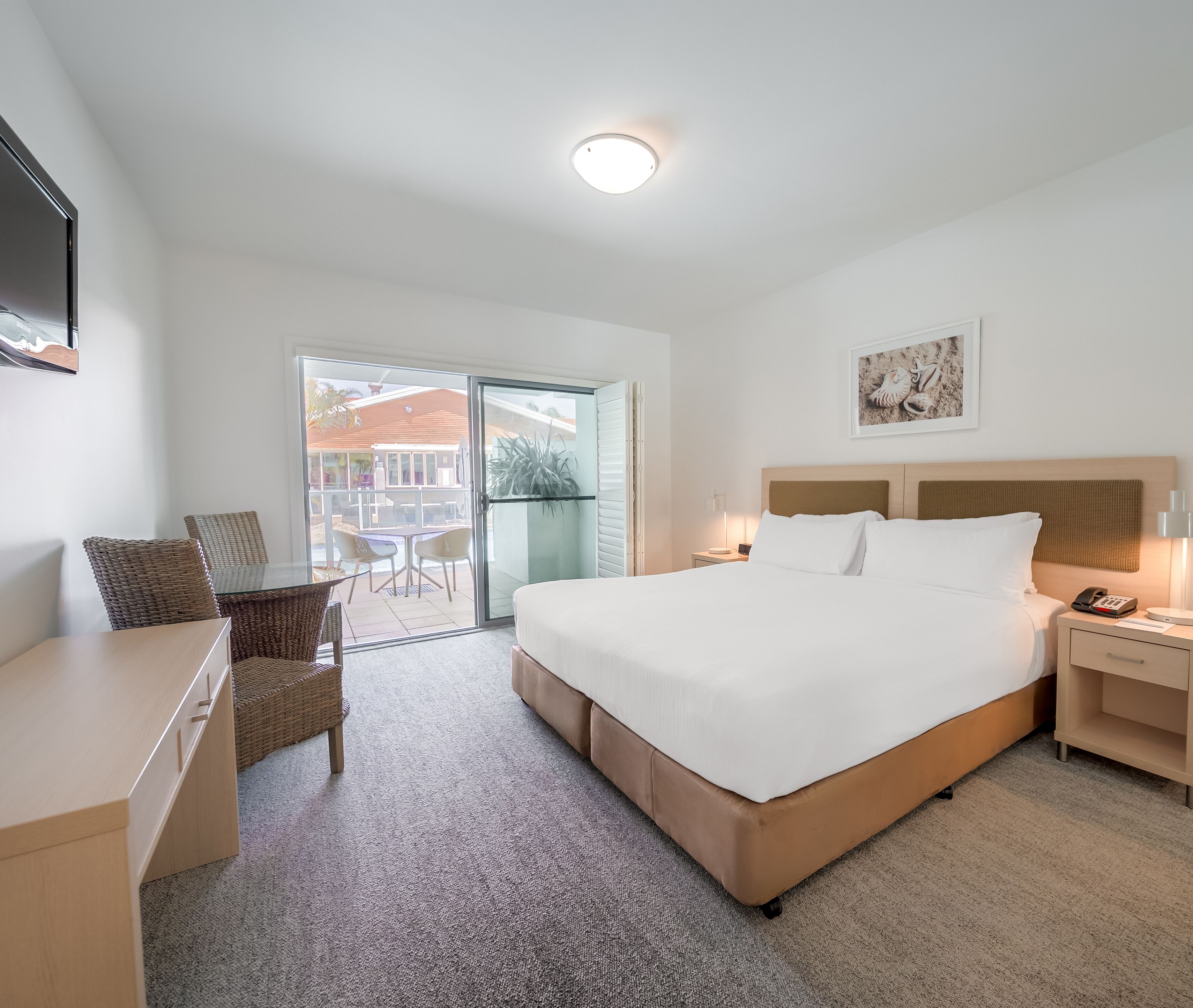 A hotel room with a bed, a desk, a chair, a nightstand, and a view of the outside.
