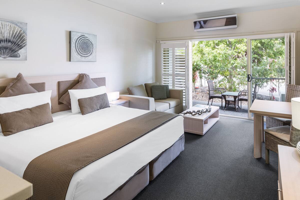 A modern hotel room with a large bed, a sofa, a dining table, and a view of the outdoors.