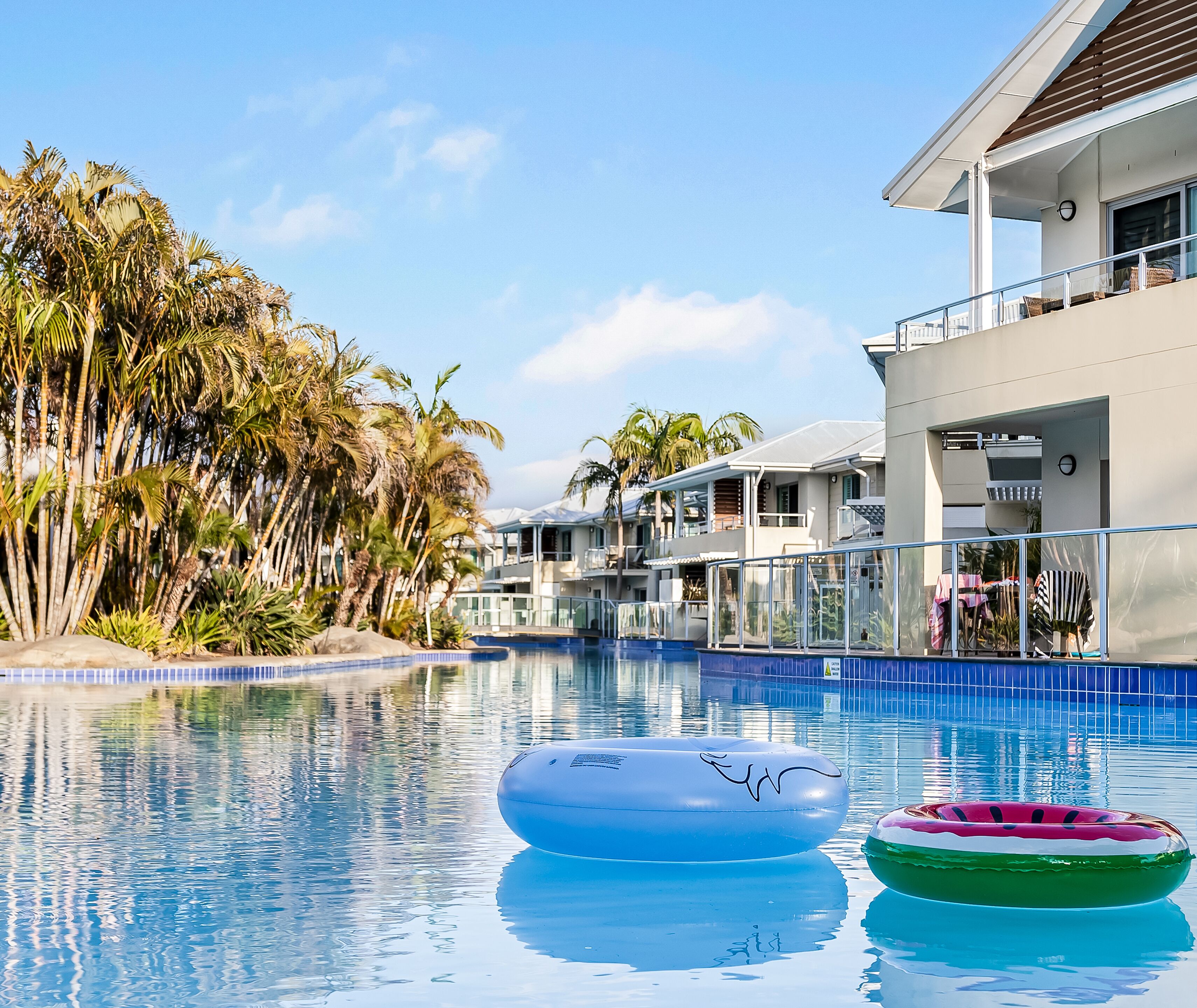 A swimming pool with two inflatable rings, surrounded by palm trees and modern buildings.