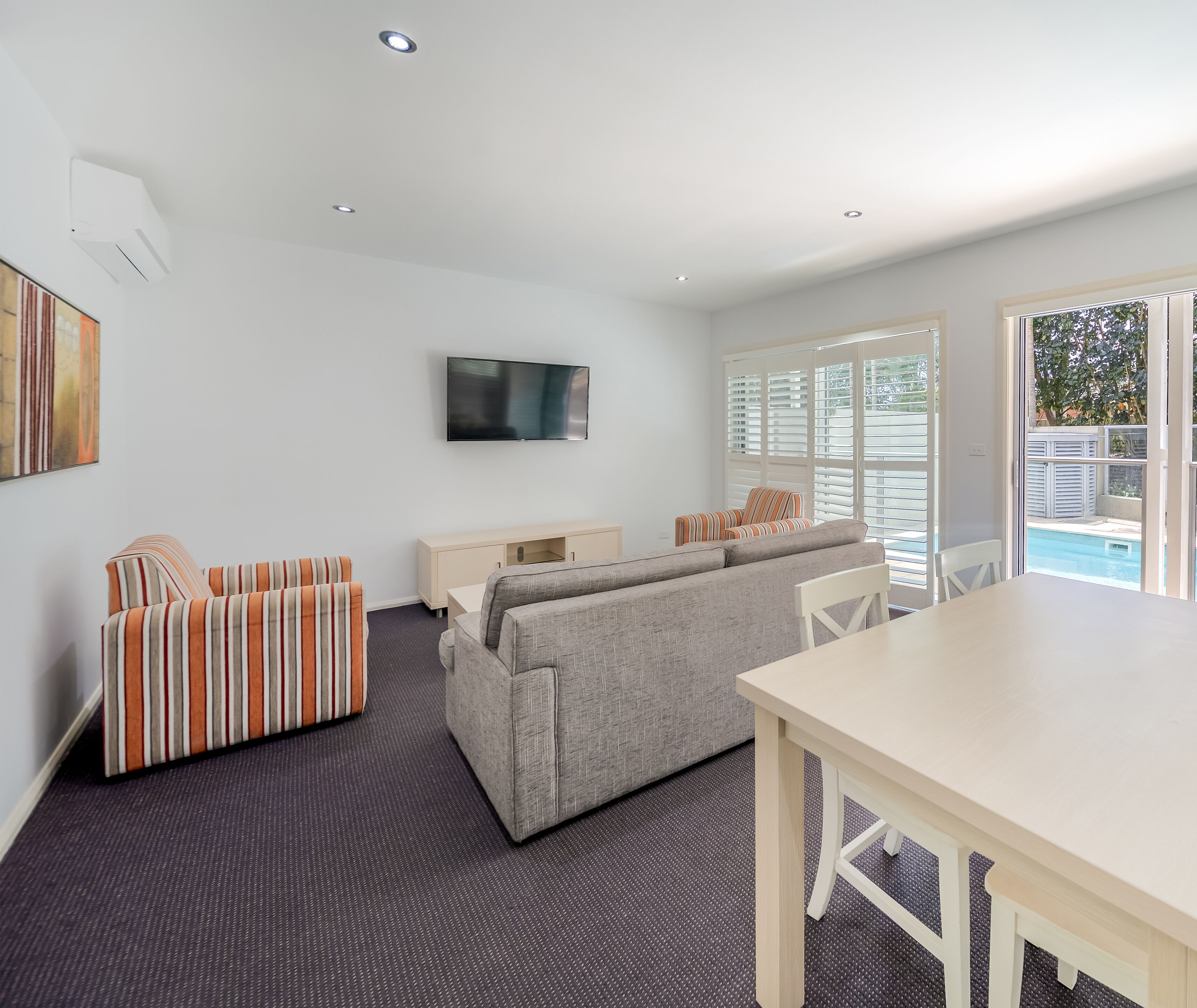 Una sala de estar moderna con un sofá gris, sillones a rayas, un televisor de pantalla plana y vista a una piscina.