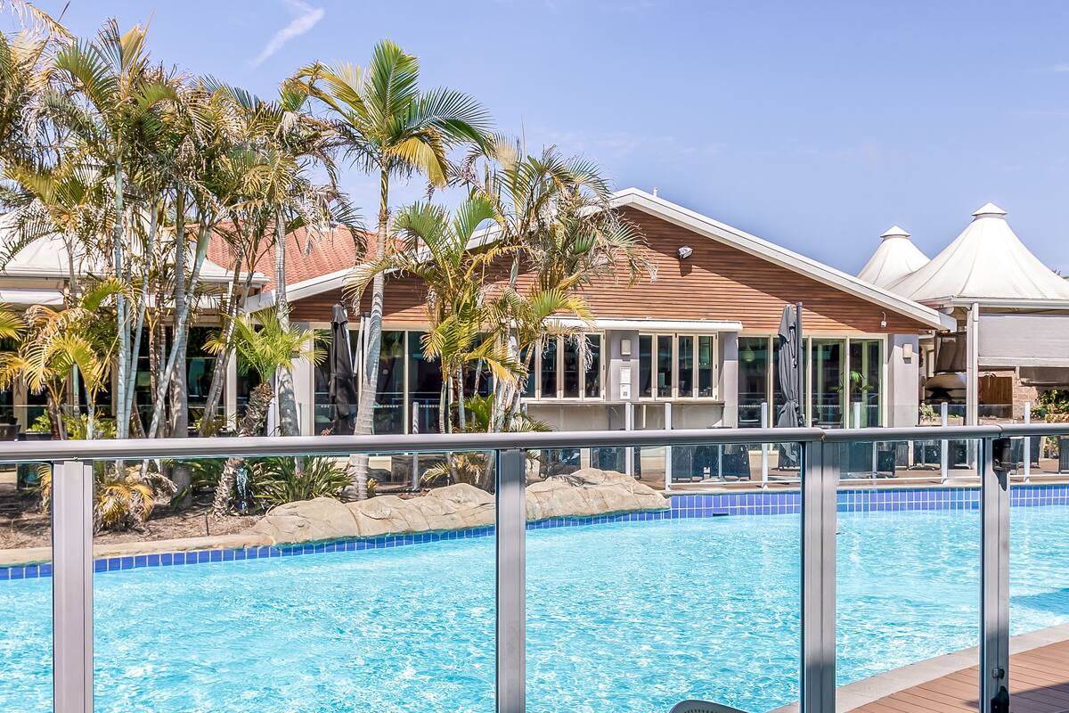A swimming pool with a clear blue water, surrounded by palm trees and a building with a tiled roof.