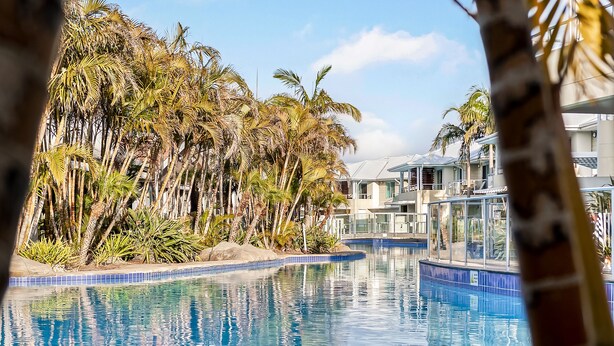 A swimming pool with clear blue water, surrounded by palm trees and a building with balconies.