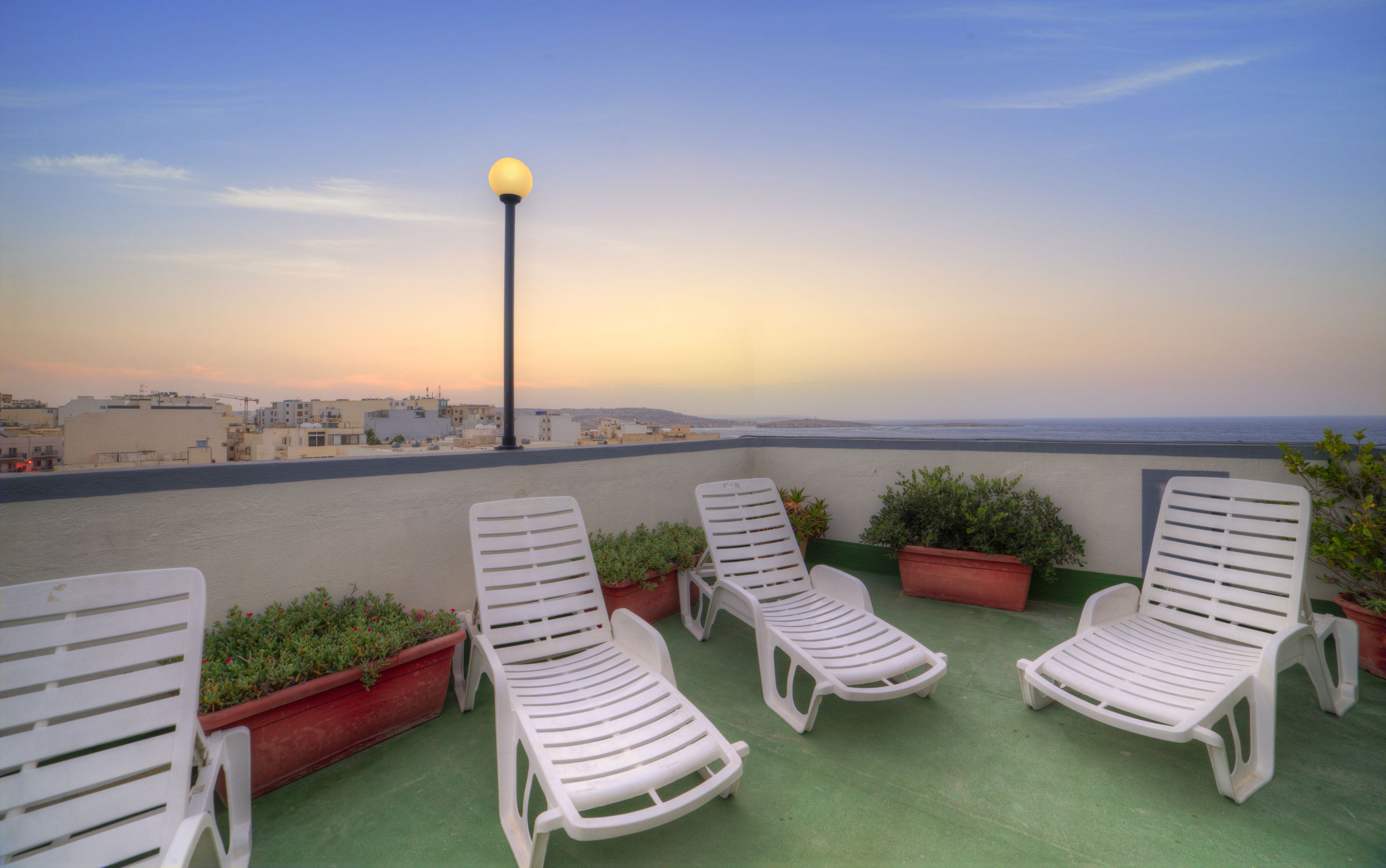 A rooftop terrace with white plastic lounge chairs, potted plants, and a street lamp.
