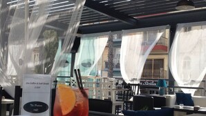 A table with a drink and a menu, under a covered outdoor seating area with a view of buildings. - Hotel Primera (St. Paul's Bay)