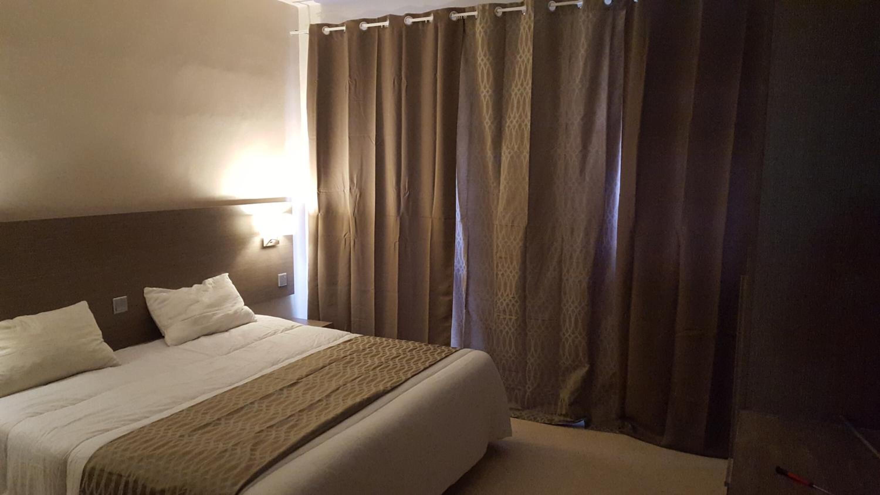 A hotel room with a large bed, a headboard, a bedside table, a lamp, and a window with curtains.