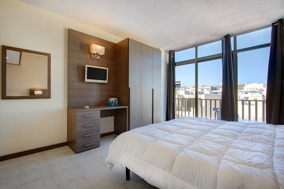 A hotel room with a bed, a desk, a TV, and a large window with a city view.
