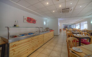 Una cafetería con mesas y sillas de madera, una barra de buffet con distintos alimentos y un cuadro en la pared.