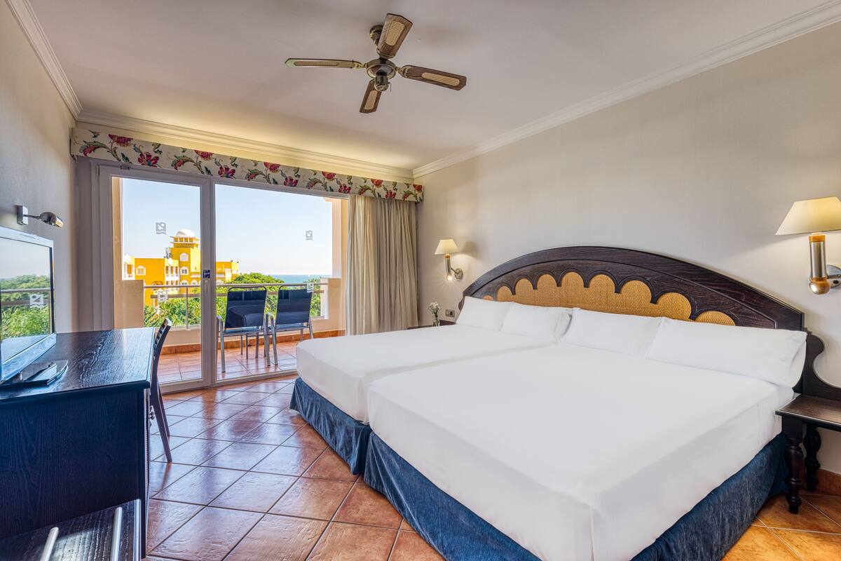 A hotel room with a large bed, a desk, a chair, a ceiling fan, and a view of buildings through the window.