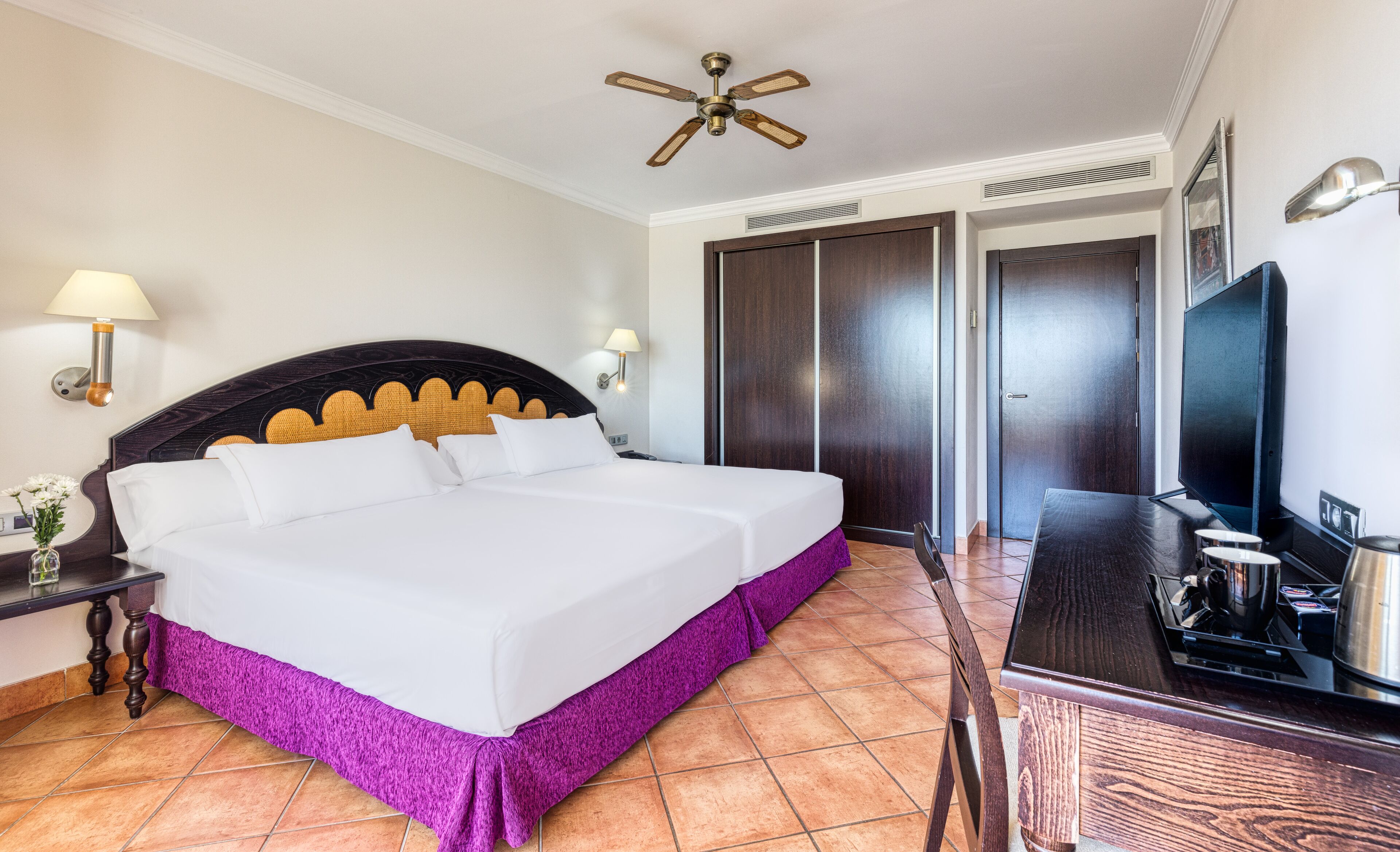 Una habitación de hotel con cama, escritorio, televisor y ventilador de techo.