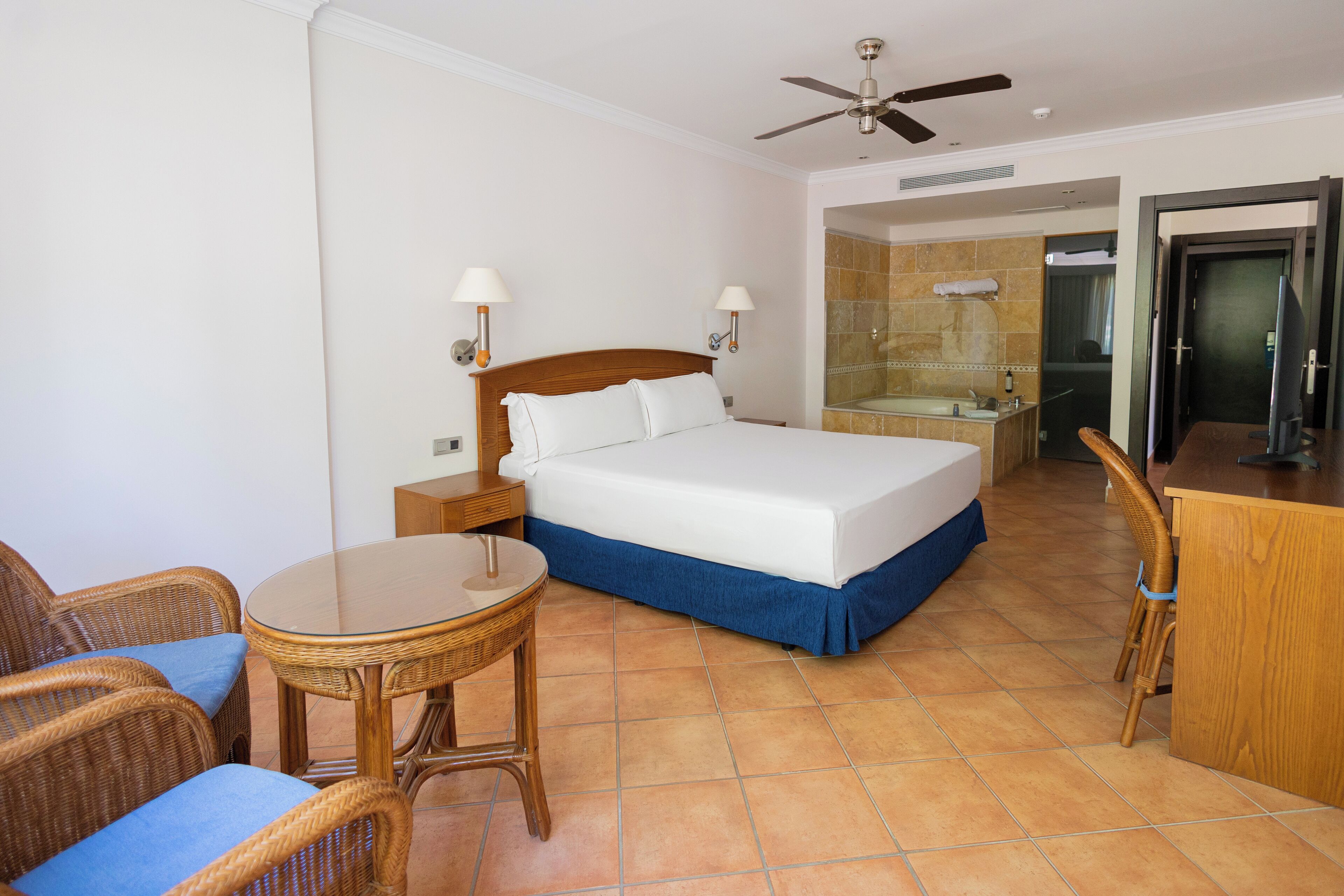 A hotel room with a bed, a small table, wicker chairs, a desk, and a bathroom visible through an open door.