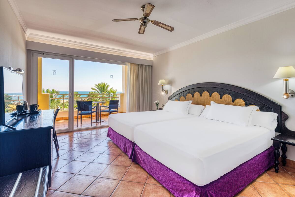 A hotel room with a large bed, a desk, a chair, a ceiling fan, a nightstand, and a view of the ocean through a sliding door.