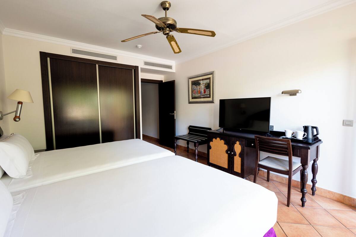A hotel room with a bed, a desk with a chair, a television, and a ceiling fan.