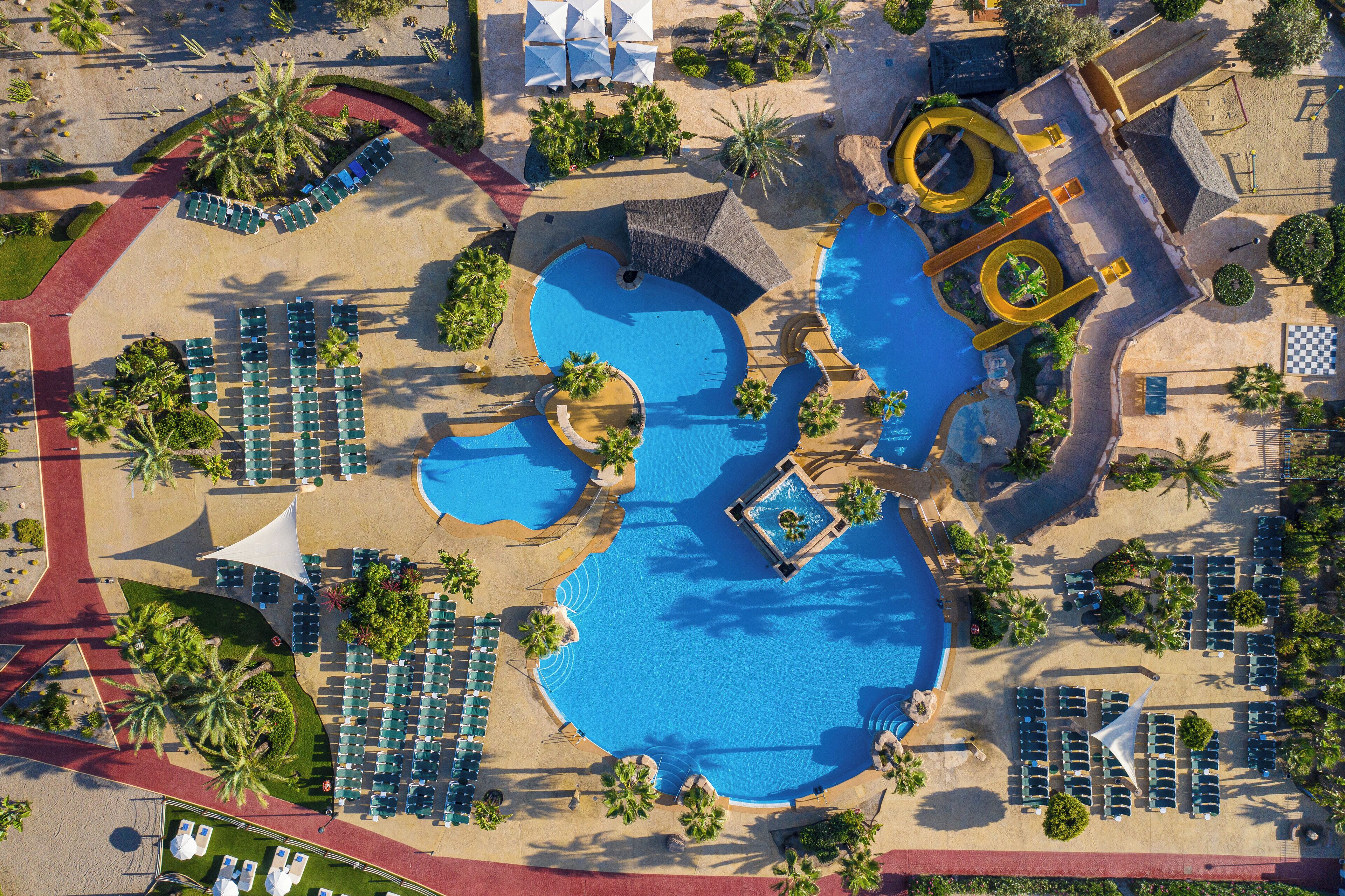 Vista aérea de un complejo turístico con varias piscinas, tumbonas y palmeras.