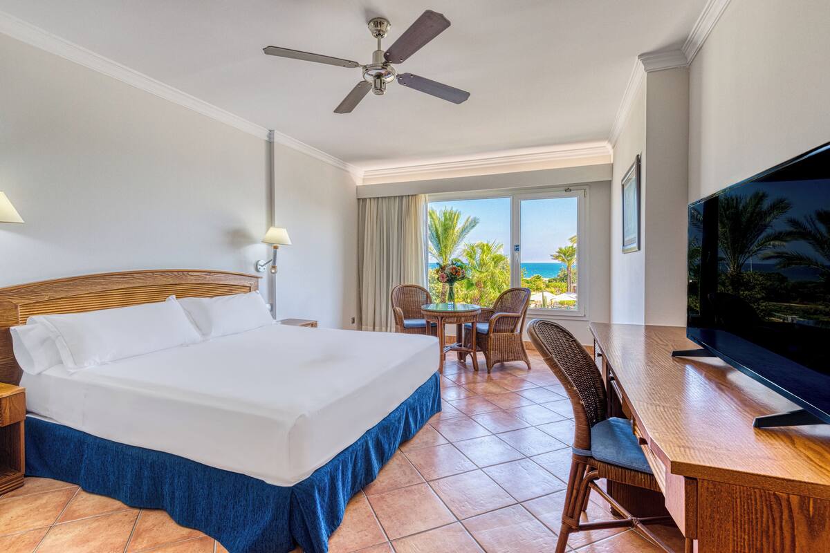 A hotel room with a bed, a TV, a ceiling fan, a balcony with a view of the ocean, and a dining area with chairs and a table.