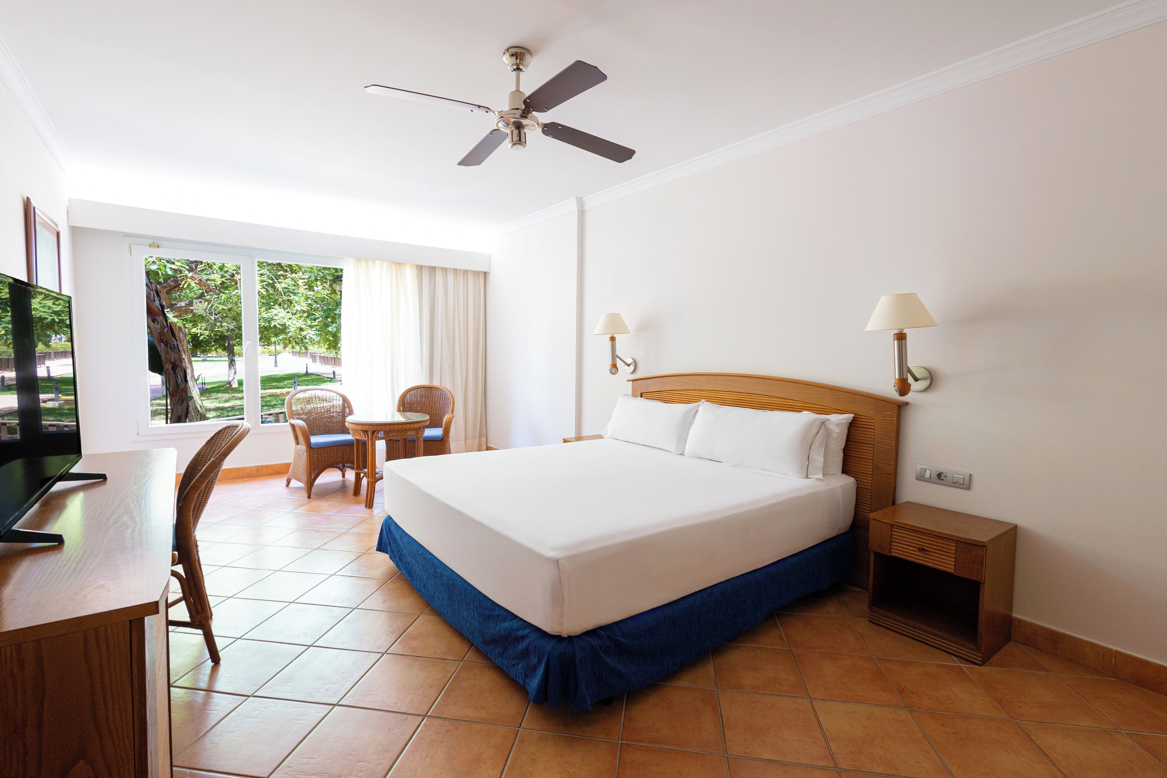A hotel room with a bed, a desk, a chair, a television, a ceiling fan, and a window with curtains.