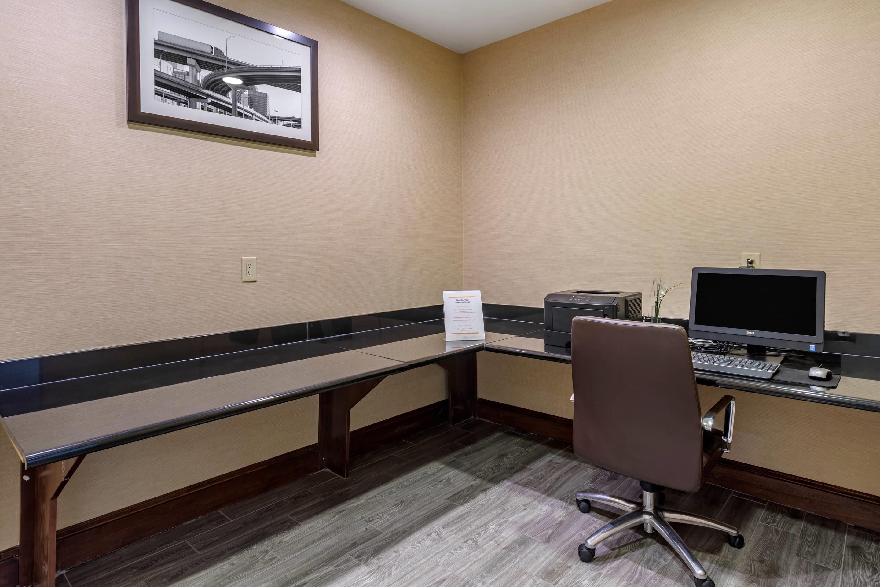 A hotel office with a desk, computer, chair, and a framed picture on the wall.