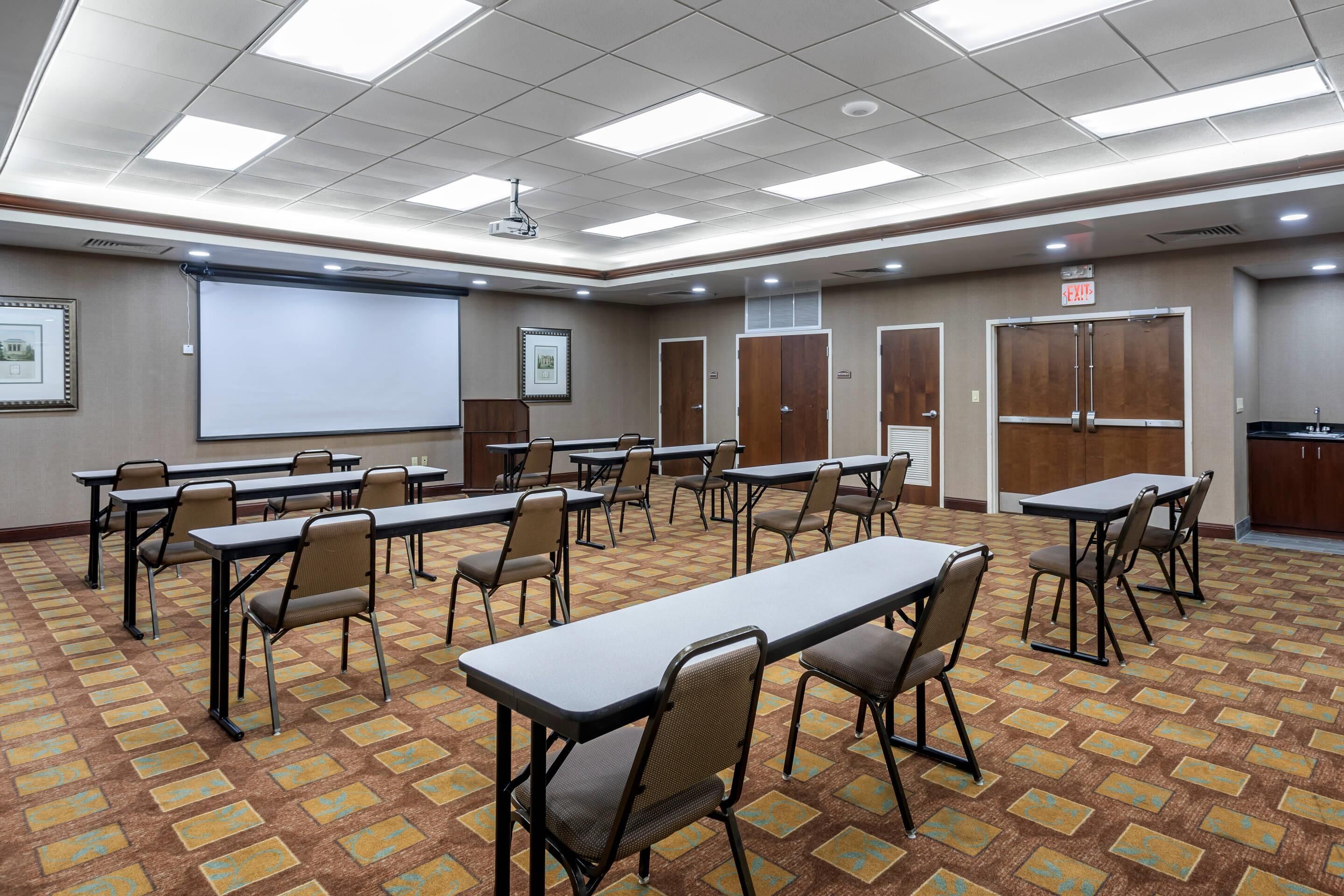 A conference room with tables, chairs, a projector screen, and a carpeted floor.