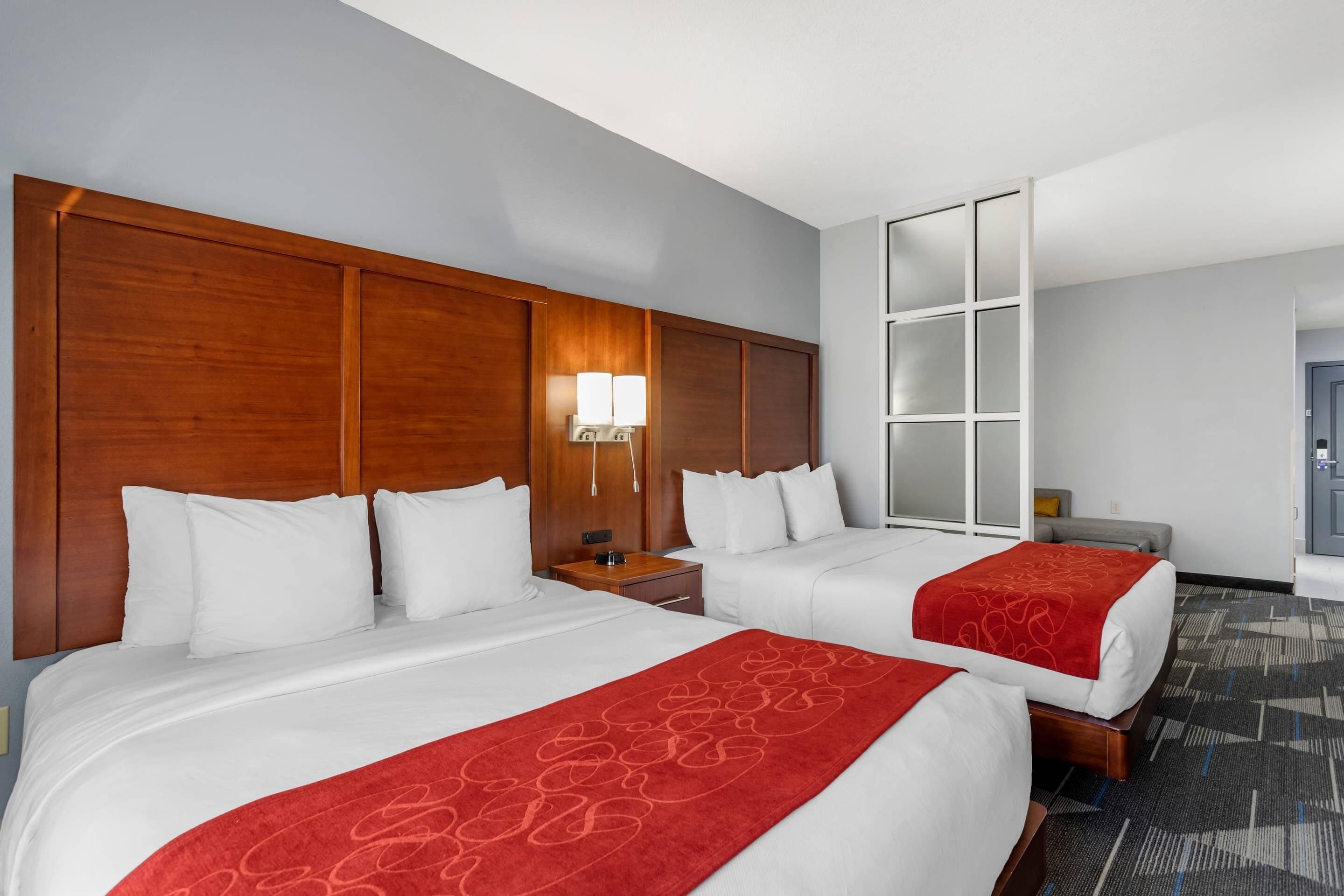 Habitación de hotel con dos camas, cabecera de madera, mesita de noche con lámpara y un ventanal grande.