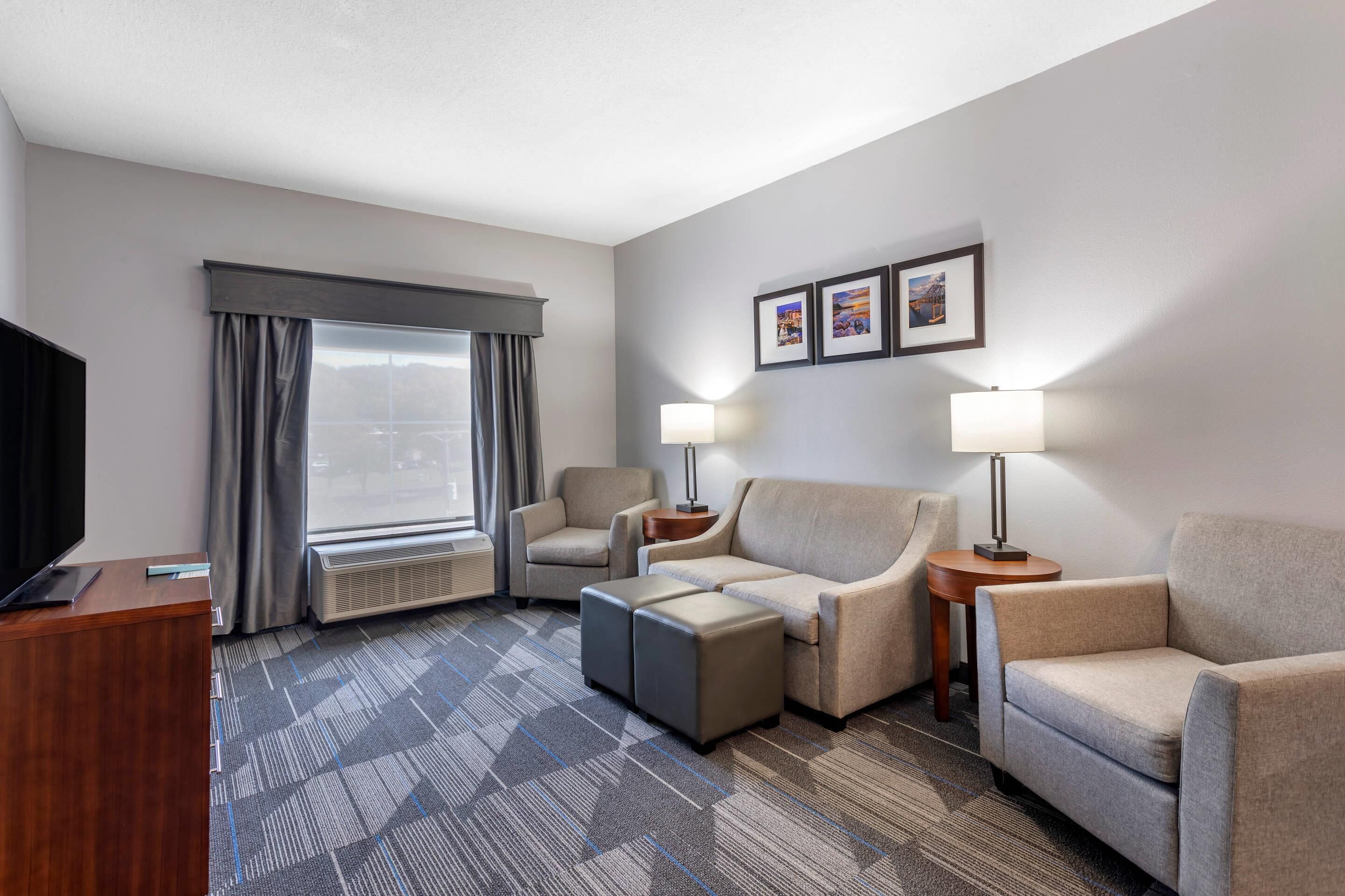 A hotel room with a flat-screen TV, a sofa, two armchairs, a coffee table, a desk, and a window with curtains.