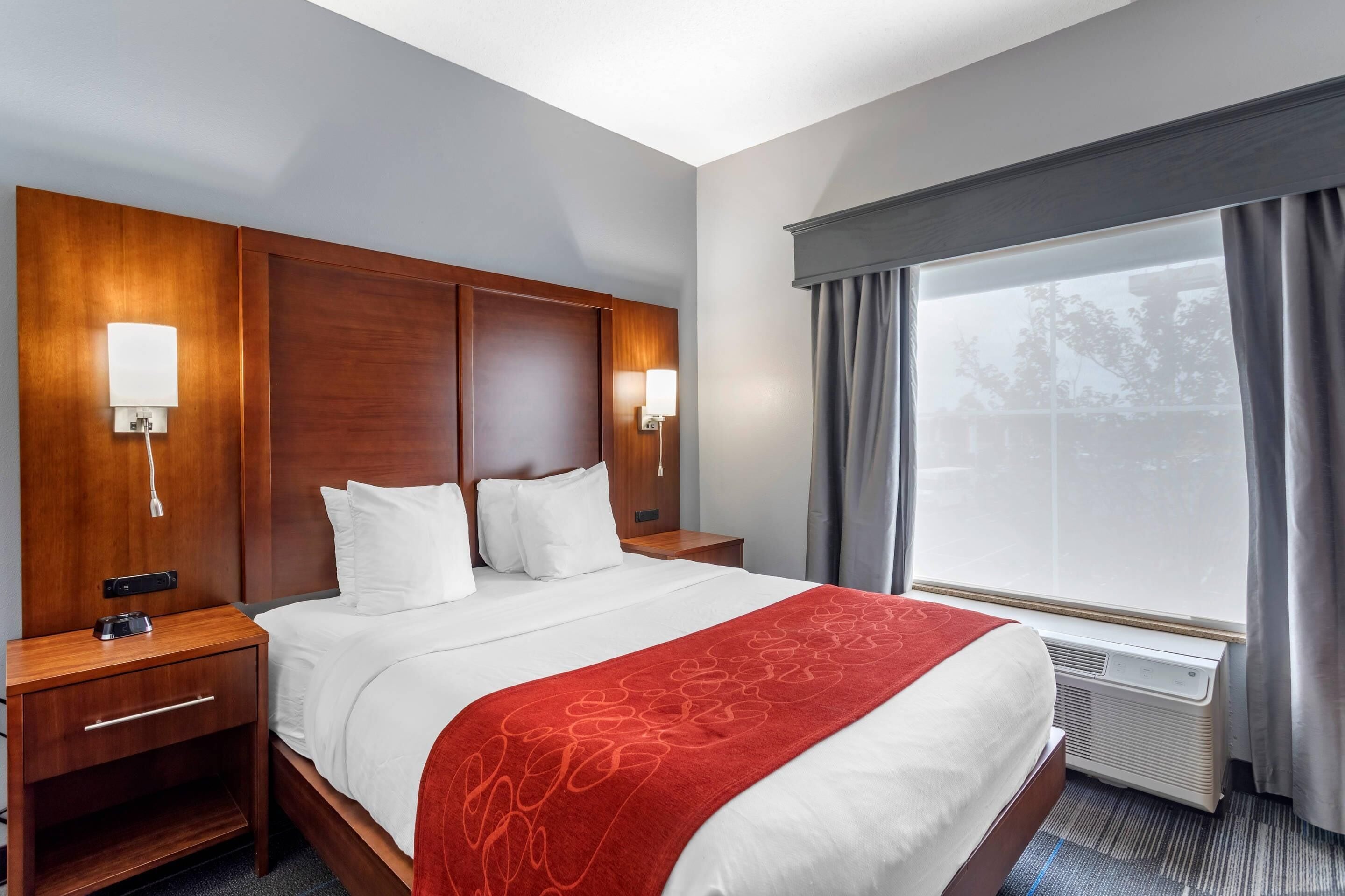 Une chambre d’hôtel avec un grand lit, une tête de lit en bois, deux lampes de chevet, une fenêtre avec des rideaux et un appareil de climatisation.