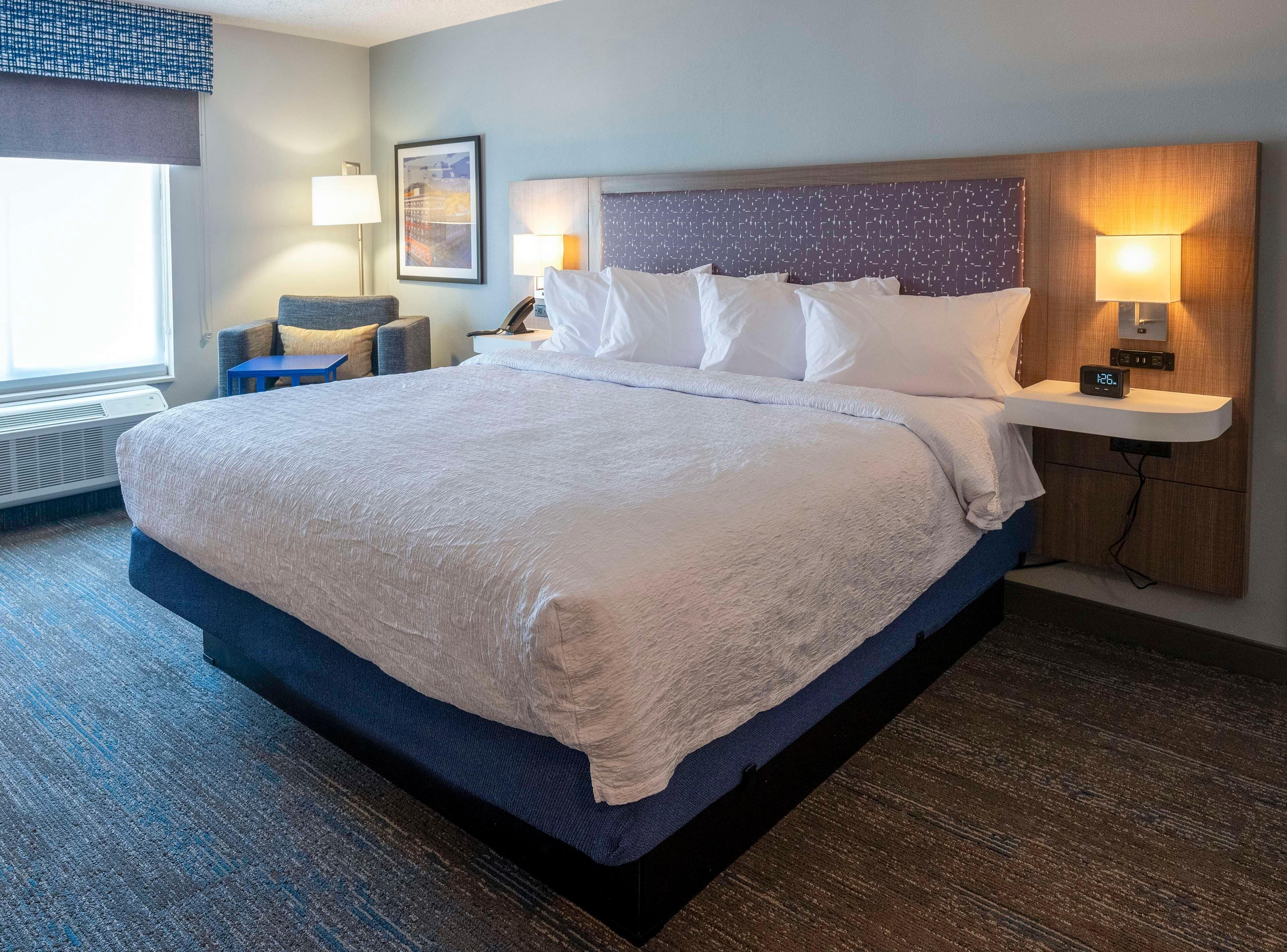 A hotel room with a large bed, bedside lamps, a small table, and a wall-mounted clock.