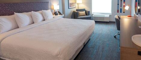 A hotel room with a large bed, a desk, a chair, a lamp, and a window with blinds.