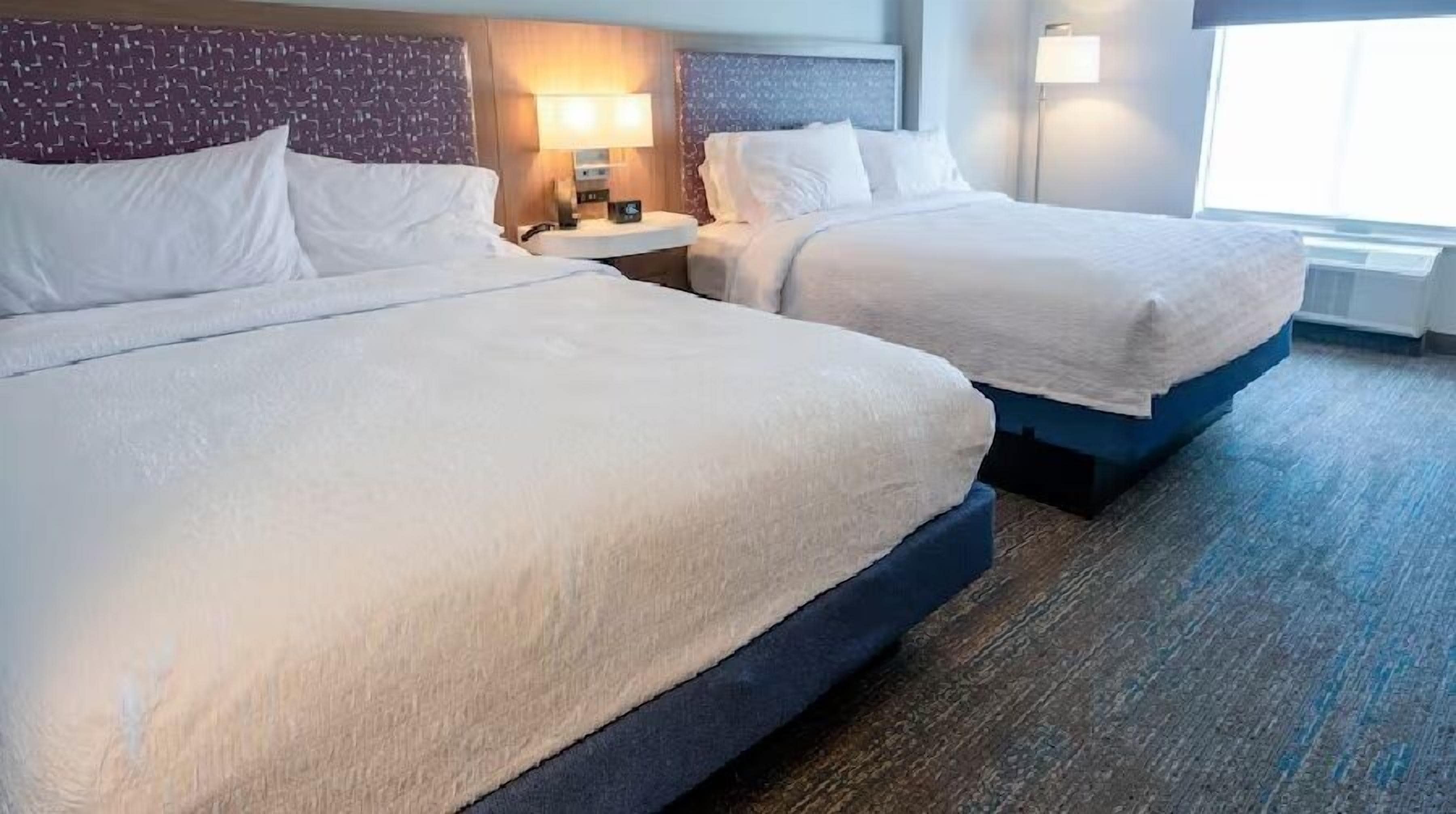 Two adjacent beds with headboards, a nightstand with a lamp, and a window with blinds.
