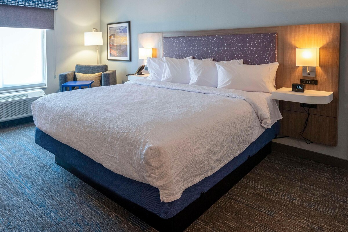 Habitación de hotel con una cama grande, lámparas de noche, una mesita y un reloj en la pared.