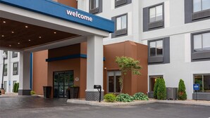 A modern hotel entrance with a 'Welcome' sign, a covered entrance area, and a landscaped front yard with plants.