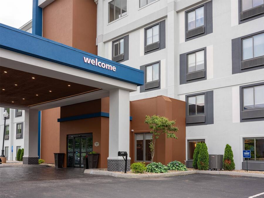 A modern hotel entrance with a 'Welcome' sign, a covered entrance area, and a landscaped front yard with plants.