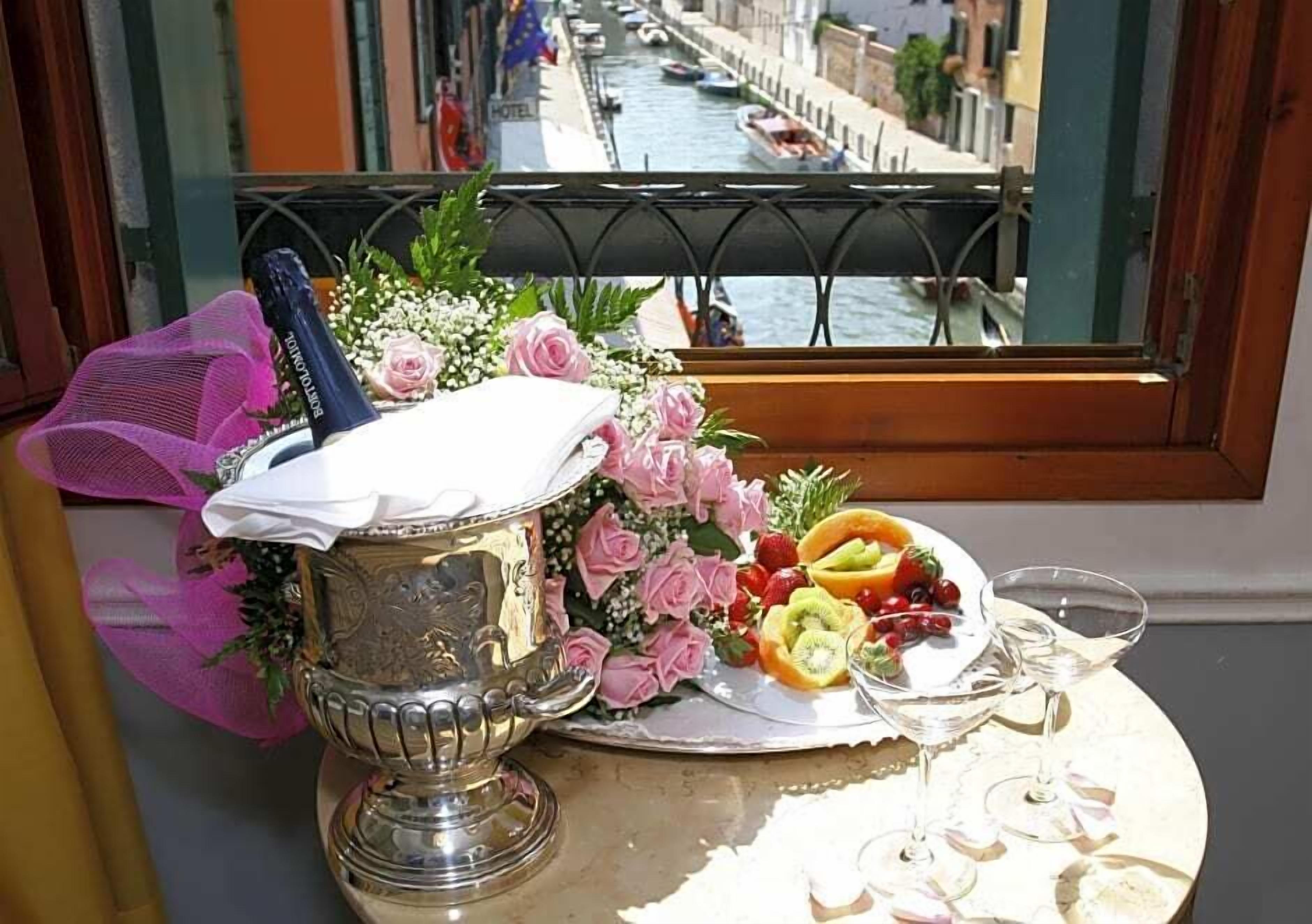 Una mesa con un candelabro de plata, un plato de fruta y copas de vino, frente a una ventana que da a un canal y barcos.