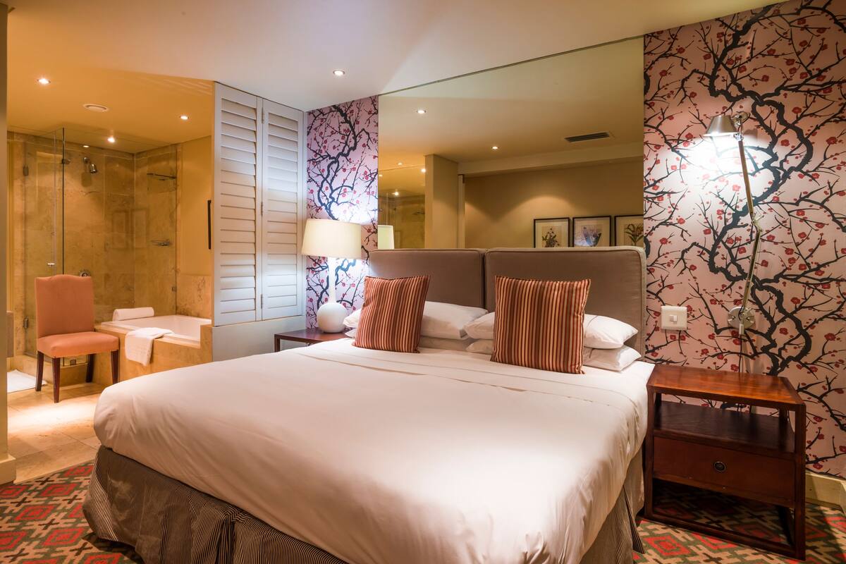 A hotel room with a large bed, a sitting area with a chair and a small table, a shower area, and a mirror with a floral wallpaper.