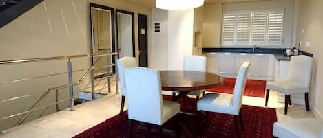 A dining area with a round table and four chairs, a staircase with a metal railing, and a kitchen with white cabinets and a window with blinds.